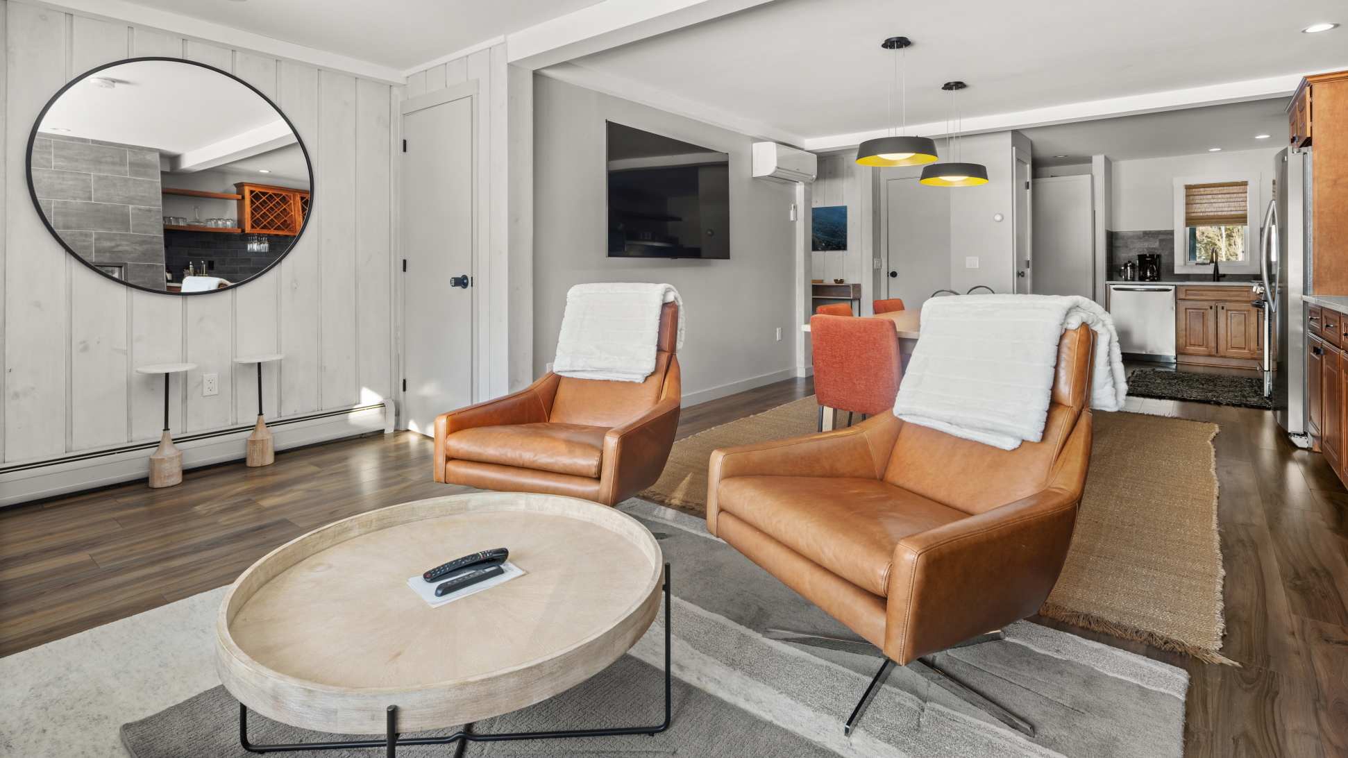 Wide view of the open-concept living area, showing two brown leather swivel armchairs facing a round wooden coffee table. A large round mirror hangs on a shiplap accent wall, and a wall-mounted TV is visible above.