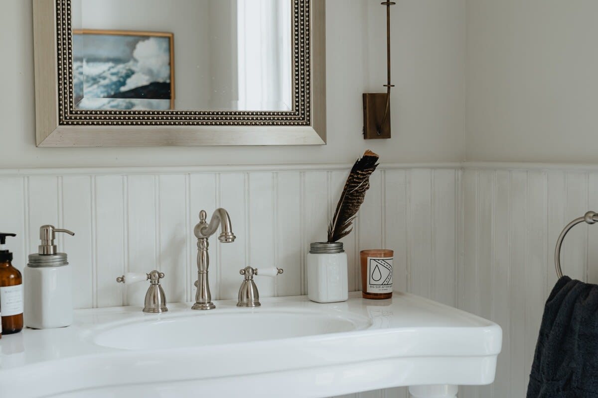 A close-up of a bathroom sink area, showcasing a white pedestal sink with brushed nickel fixtures and a silver framed mirror. Decorative items like a feather, candle, and soap dispenser adorn the counter, set against white beadboard wainscoting.