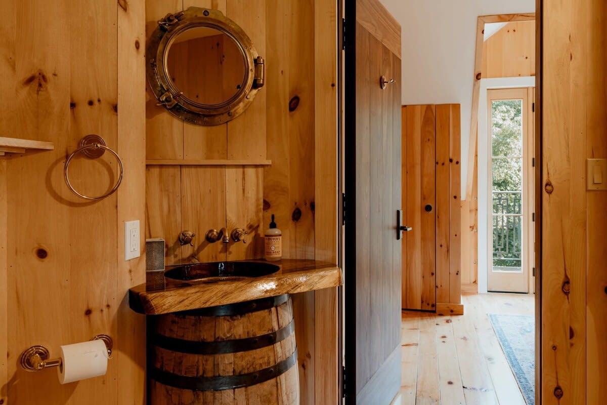 Rustic bathroom featuring light wood paneling, a unique barrel vanity with a dark basin, and a brass porthole mirror, creating a nautical-inspired aesthetic.
