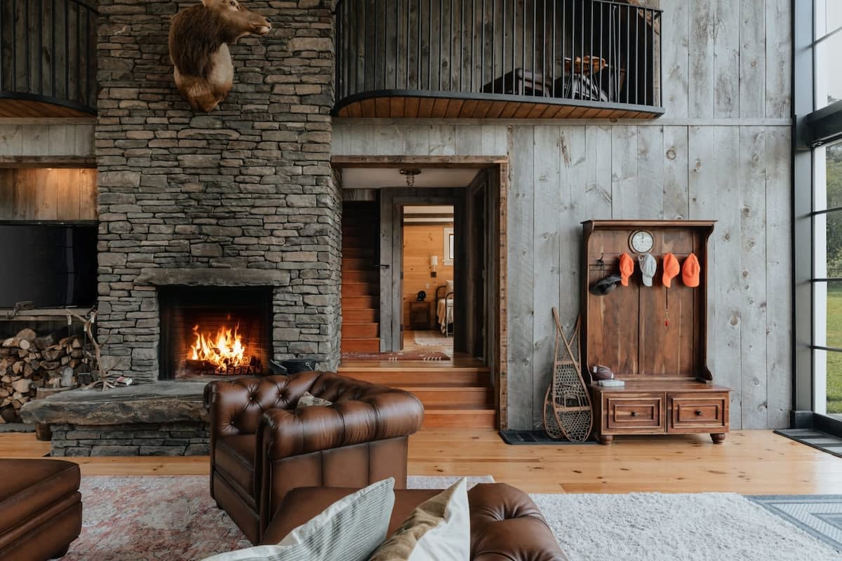 A close-up view of the grand living room, highlighting the large stone fireplace with a roaring fire and stacked firewood. A brown leather armchair is visible, along with a rustic wooden hall tree and a glimpse of the upper-level balcony.