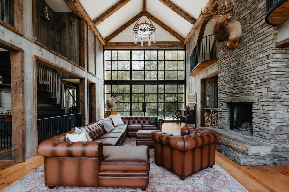 A grand living room with a vaulted ceiling, exposed wooden beams, and a large stone fireplace. Brown leather Chesterfield sofas and armchairs are arranged on a patterned rug, facing a wall of tall windows overlooking a wooded landscape.