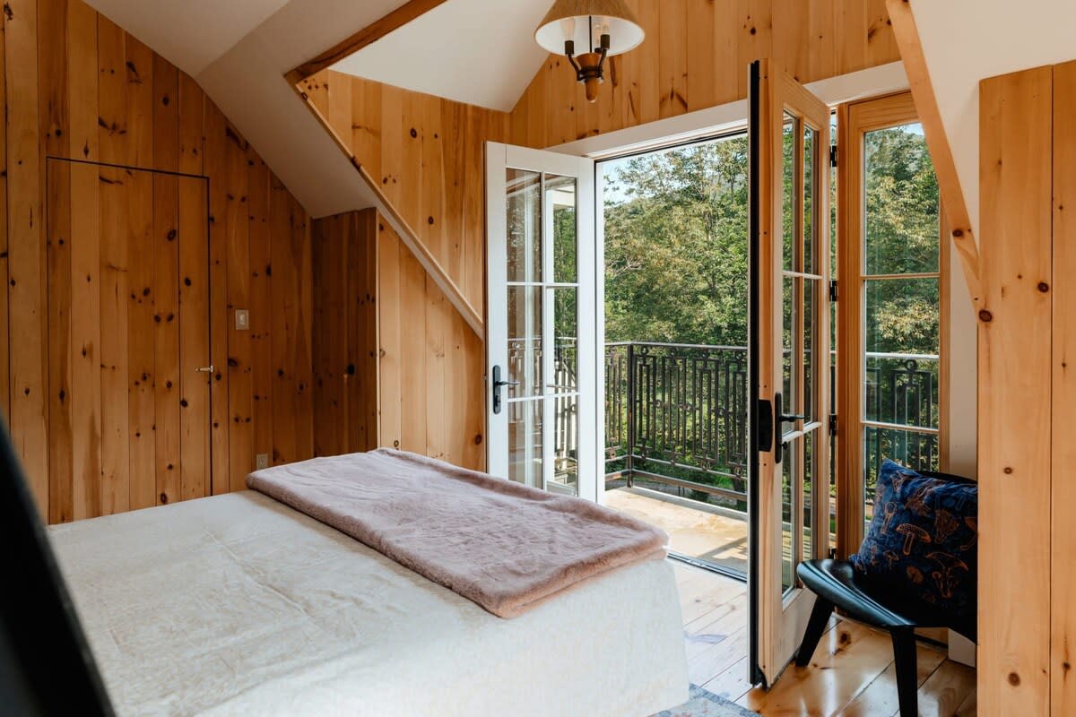 Bright bedroom with light wood paneling, a bed with a pink throw, and French doors opening to a balcony with a view of lush trees and natural surroundings.
