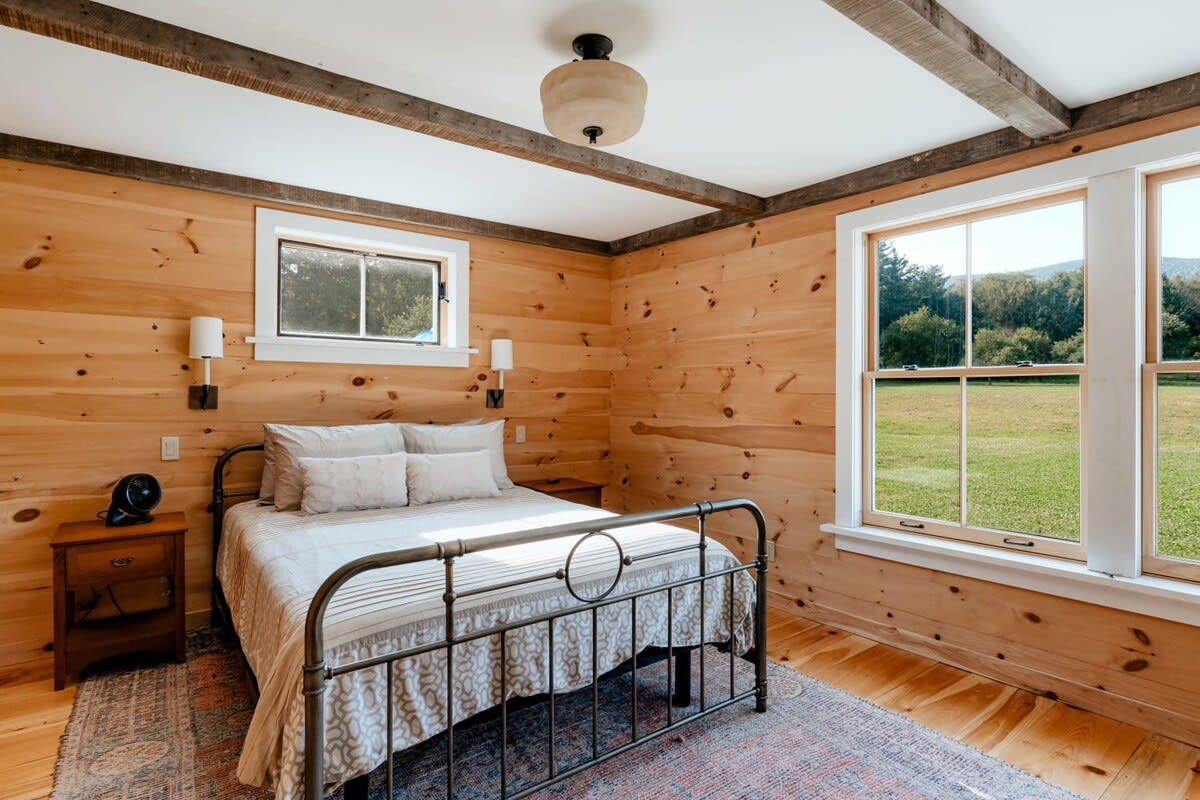 A rustic bedroom with light wood plank walls and hardwood floors, featuring a wrought iron queen-sized bed with white bedding. Two large windows offer views of a grassy field, and exposed wooden beams accent the ceiling.