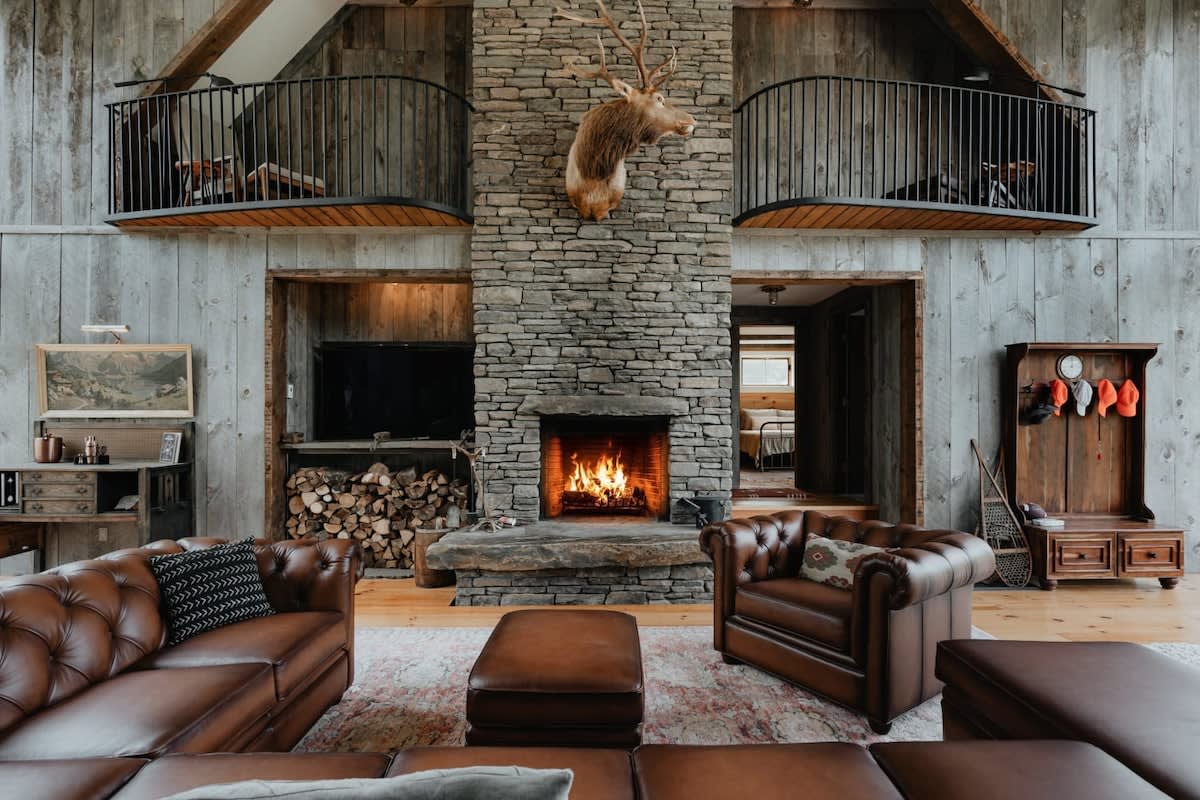 Cozy rustic living room with a large stone fireplace, mounted deer head, and two brown leather Chesterfield sofas. Two curved balconies with black railings overlook the space from above.