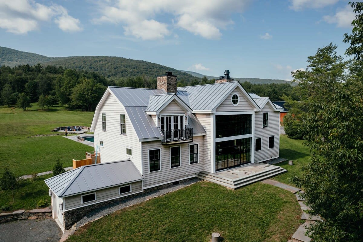 Side view of the white farmhouse with a gray metal roof and multiple gables, featuring a large black-framed window facade and a small balcony. The house is set on a green lawn with mountains in the background.