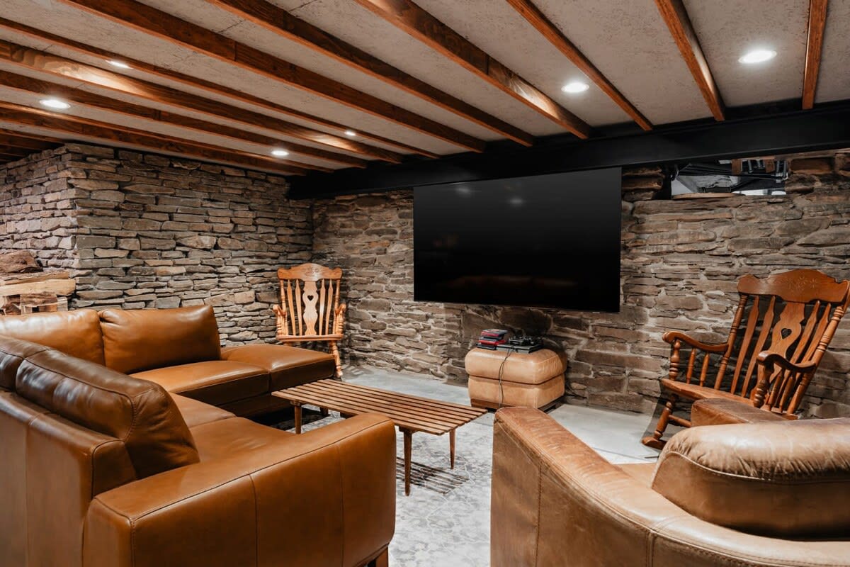 Spacious media room with rustic stone walls, exposed wooden beams on the ceiling, comfortable brown leather sofas, and a large wall-mounted television for entertainment.