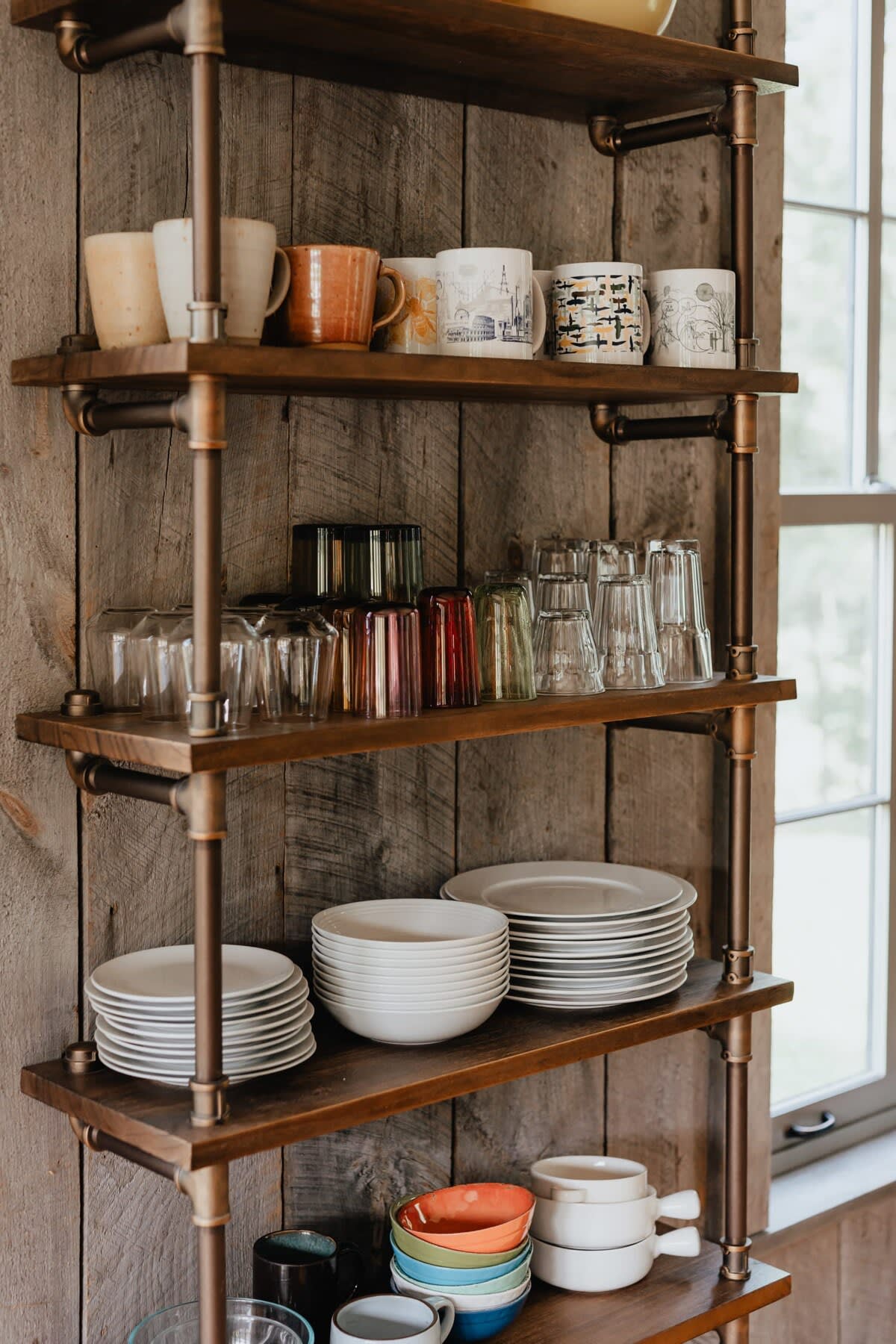 A rustic open shelving unit made of wood and metal pipes, displaying various mugs, glasses, plates, and bowls. The shelves are set against a light wood-paneled wall next to a window.