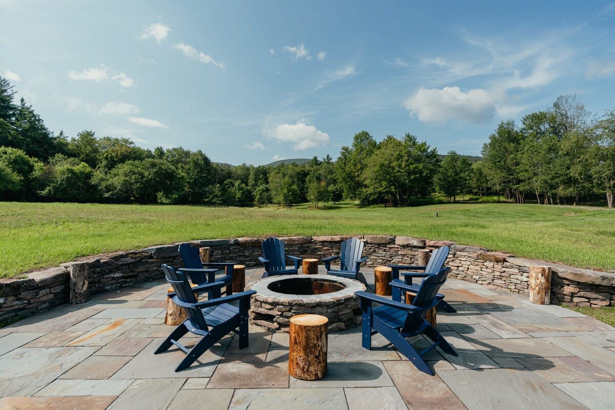 An outdoor fire pit area with a circular stone wall, surrounded by blue Adirondack chairs and tree stump side tables, set on a stone patio overlooking a vast green field and distant mountains under a blue sky.