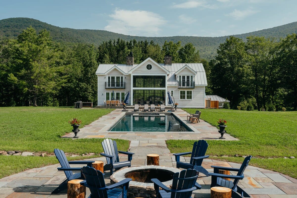 Aerial view of the white farmhouse and its rectangular swimming pool, leading to a stone fire pit area with blue Adirondack chairs. The property is surrounded by vast green lawns and mountains.