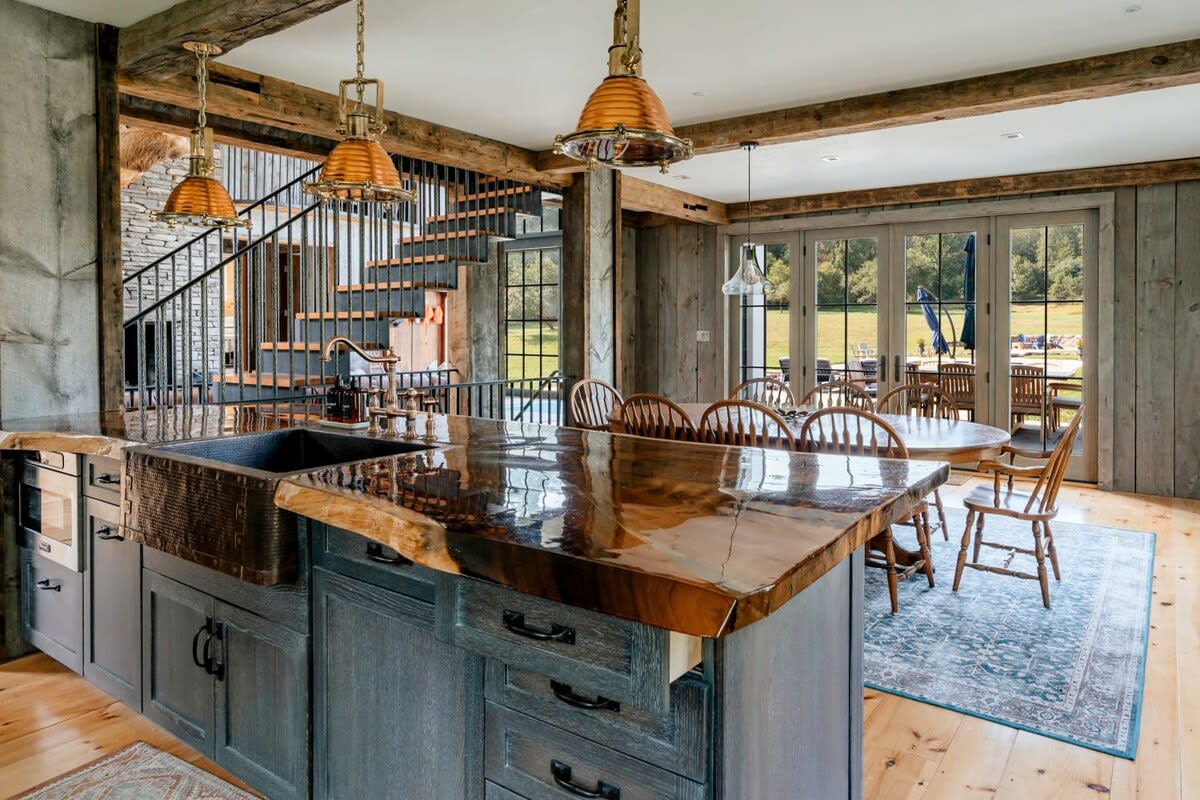A rustic-chic kitchen with a large island featuring a live-edge wooden countertop and a farmhouse sink. Exposed wooden beams, industrial pendant lights, and a view of the outdoor area through French doors complete the space.