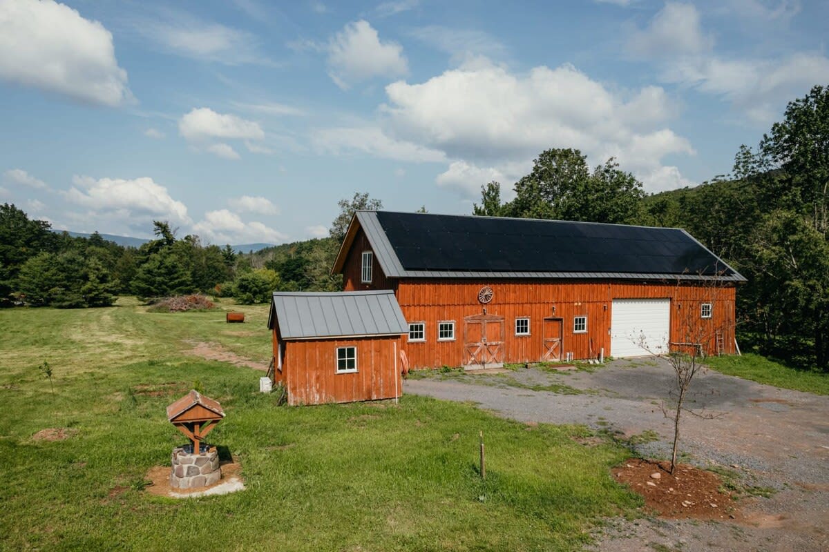 A large red barn with a black metal roof covered in solar panels stands prominently in a green field, accompanied by a smaller matching shed and a decorative stone wishing well, with forested hills in the background.