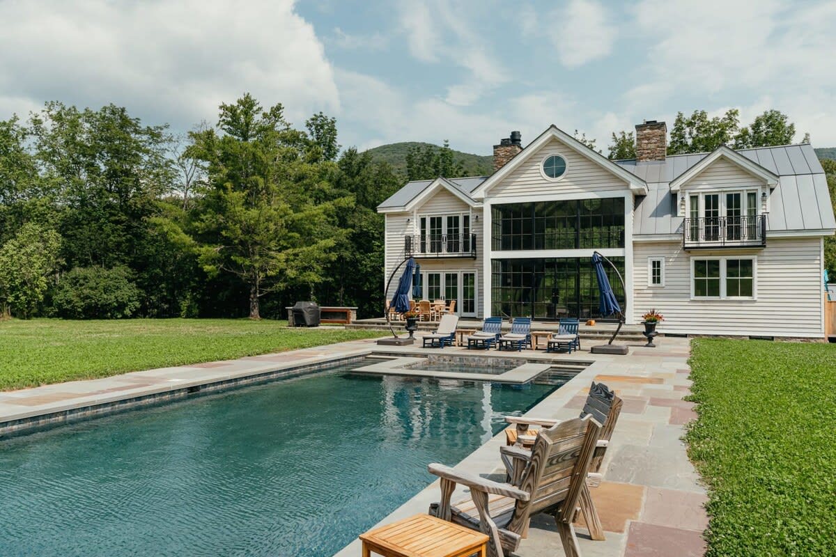 The modern farmhouse features a large rectangular swimming pool with an integrated hot tub, surrounded by a stone patio with lounge chairs and umbrellas, all set against a lush green lawn and distant mountains.
