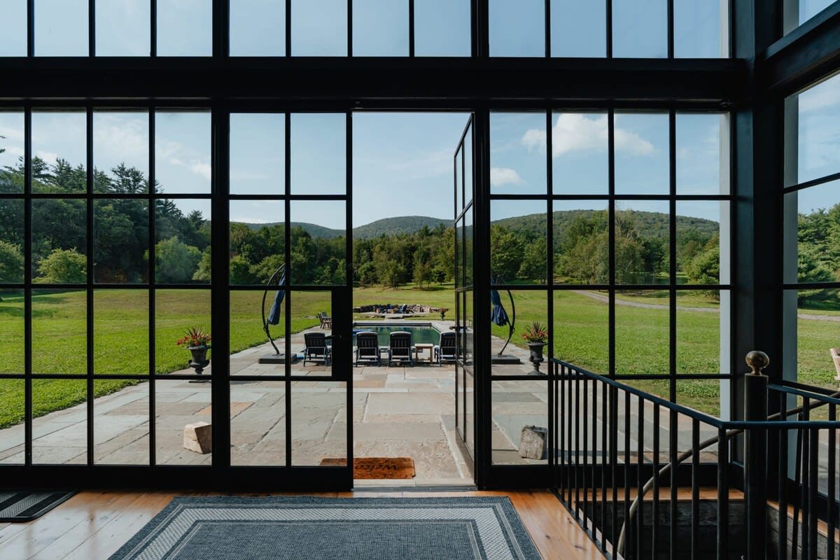 View from inside a modern farmhouse looking out through large black-framed glass doors to a bluestone patio, swimming pool, and expansive green landscape with distant mountains. A black metal railing for stairs is visible on the right.