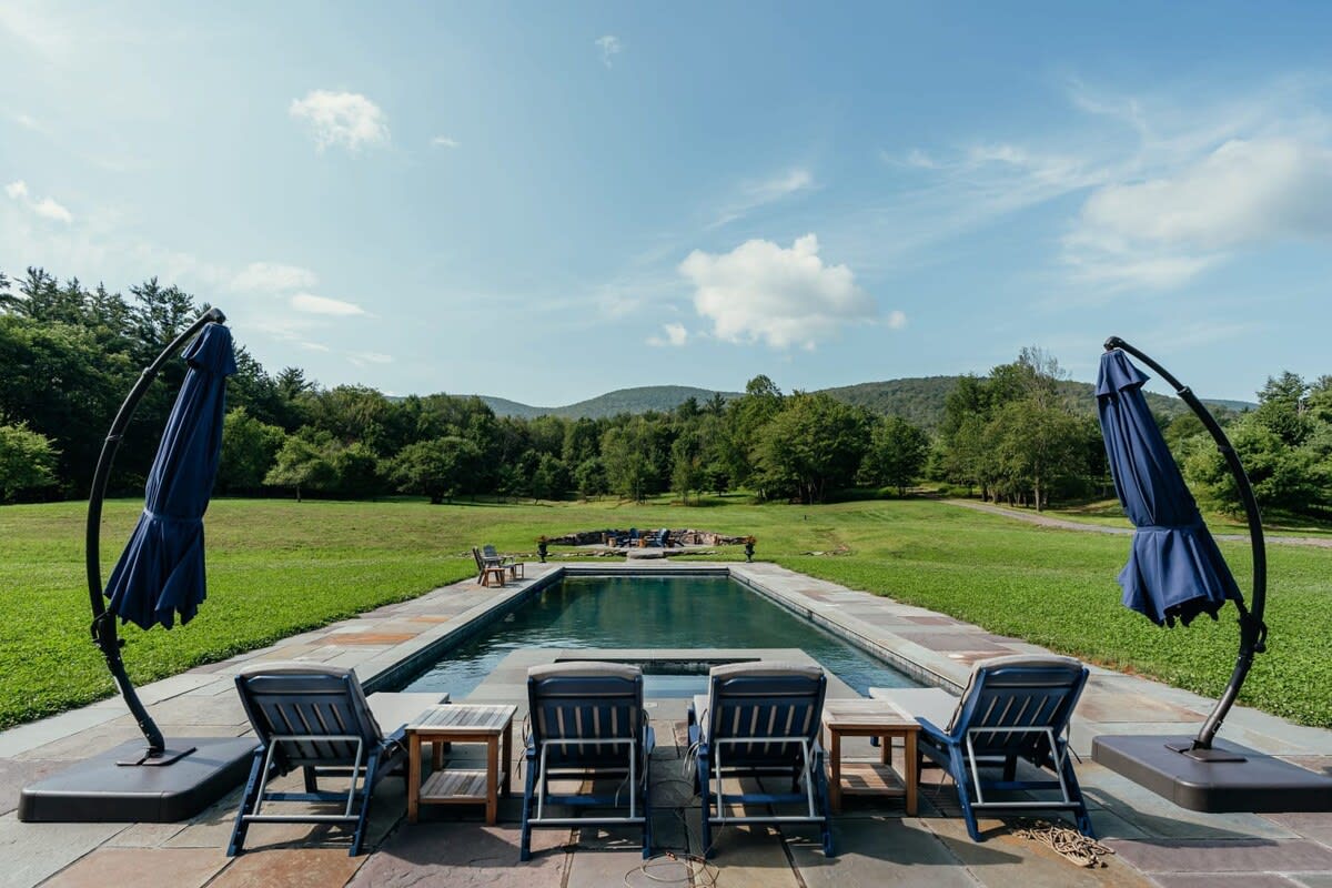 Rectangular outdoor swimming pool surrounded by a stone patio, with lounge chairs, two large blue umbrellas, and a picturesque mountain backdrop, perfect for relaxation.
