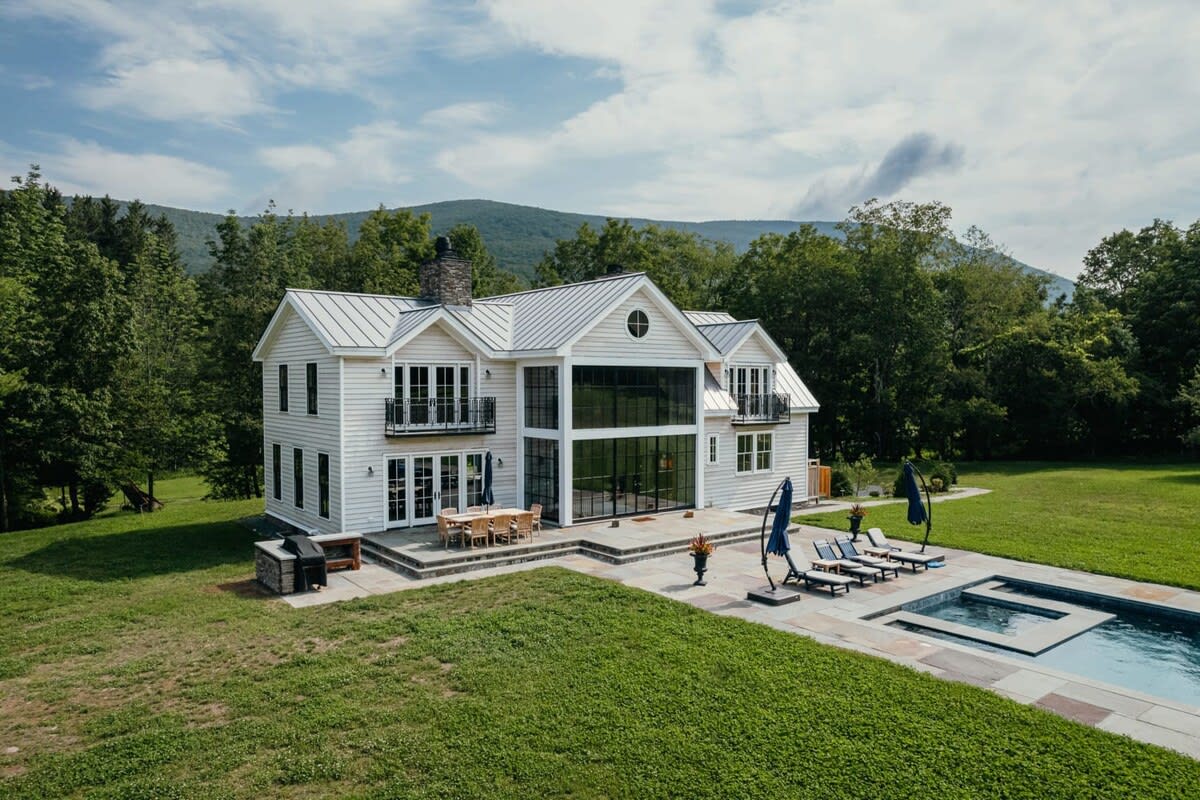 The rear of the modern farmhouse showcases a multi-level stone patio with an outdoor dining area and a BBQ station, extending towards a pool deck with lounge chairs, all framed by lush green grass and distant trees.