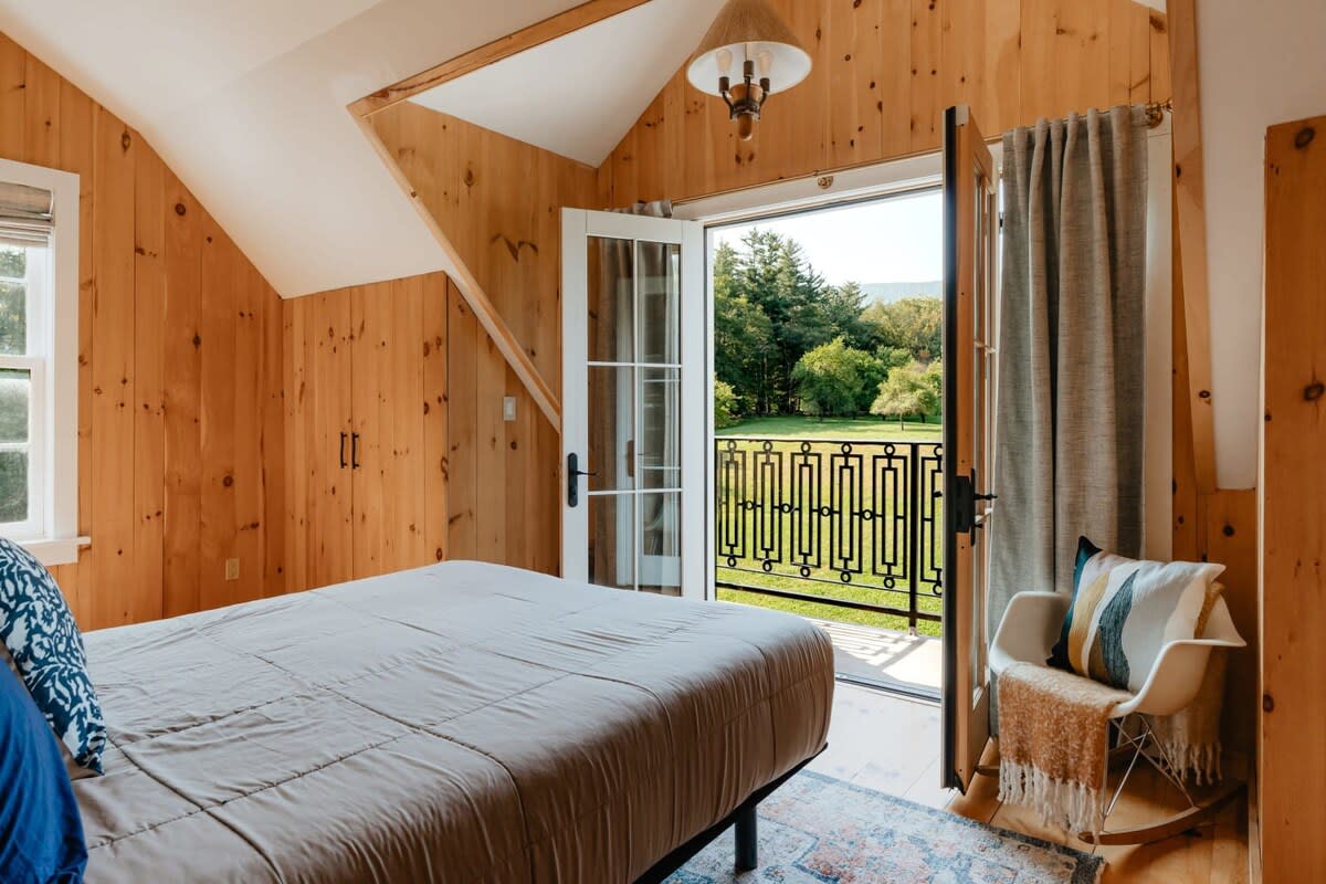 Inviting bedroom with light wood paneling, a bed with a grey duvet, and French doors opening to a balcony overlooking a lush green lawn and distant trees.