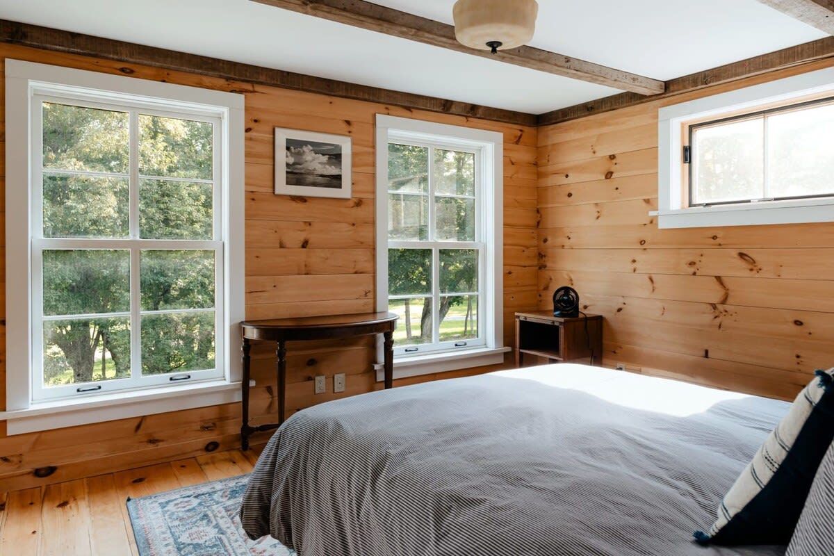 A cozy bedroom featuring light wood plank walls and floors, two large windows overlooking lush greenery, and a bed with a striped duvet. A small wooden console table sits between the windows, and a ceiling light fixture hangs from a beamed ceiling.
