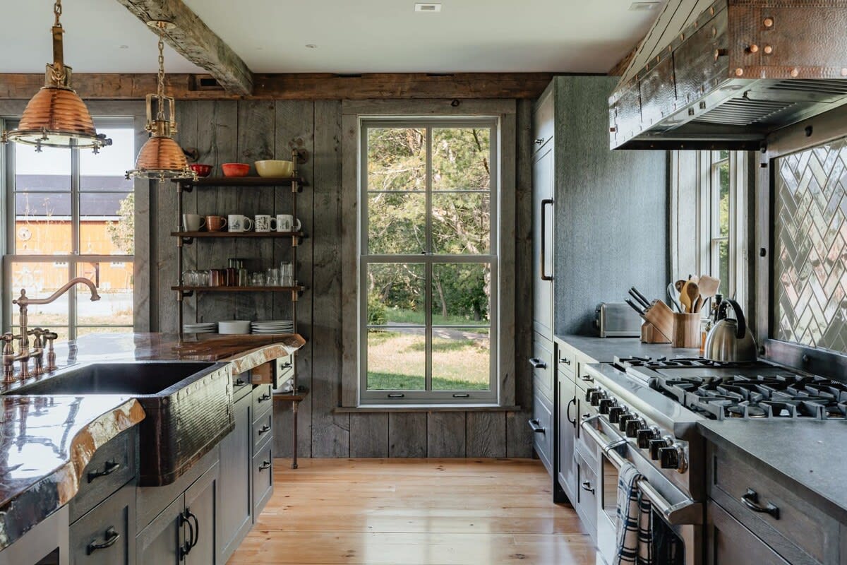 Rustic kitchen with a large farmhouse sink set in a live-edge wooden island, dark wood cabinetry, and open shelving. Copper pendant lights illuminate the space, which features wood paneling and hardwood floors.