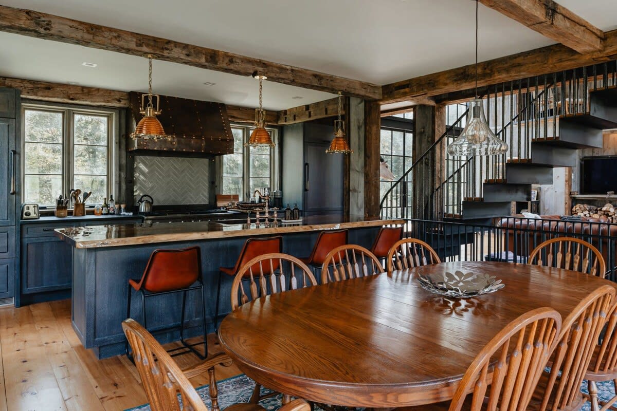 A combined kitchen and dining area featuring a large wooden dining table with spindle-back chairs, adjacent to a kitchen island with bar stools. The space has exposed wooden beams, hardwood floors, and a view of the outdoor area.