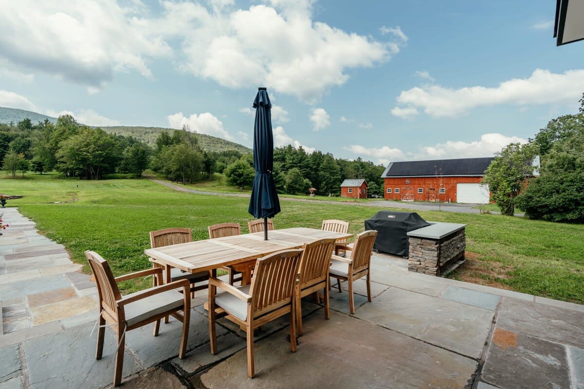 Outdoor patio with a large wooden dining table and chairs, a black umbrella, a BBQ grill, and a scenic view of a red barn and expansive green fields.