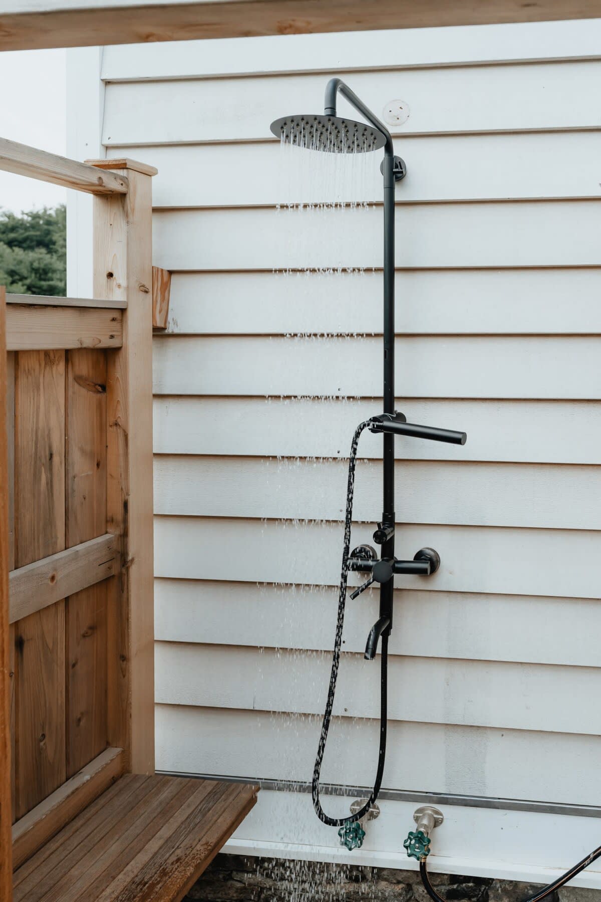 A rustic outdoor shower with black industrial-style fixtures is mounted on the white shiplap siding of the house, featuring a wooden privacy screen and slatted floor.