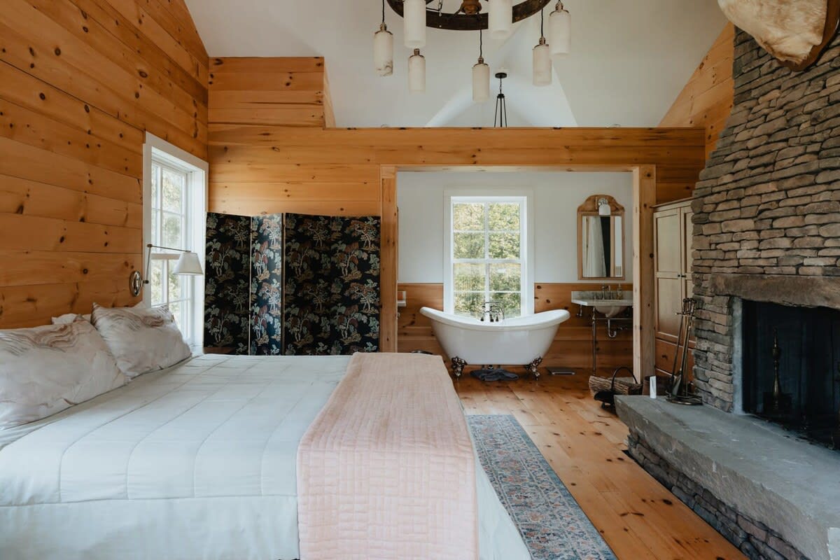 A wide shot of a master bedroom featuring warm wood plank walls and a vaulted ceiling, with a queen bed and a prominent stone fireplace. An open alcove reveals a freestanding clawfoot tub and a vanity, with large windows offering views of the outdoors.