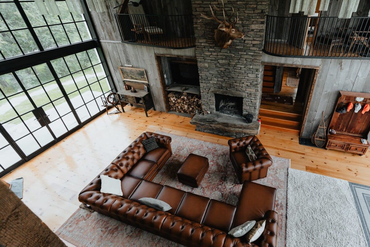 An elevated view of the spacious living room, showcasing the arrangement of brown leather sectional and armchair on a large rug. The room features a high ceiling, a stone fireplace, and a wall of windows, with a rustic wooden hall tree near the entrance.