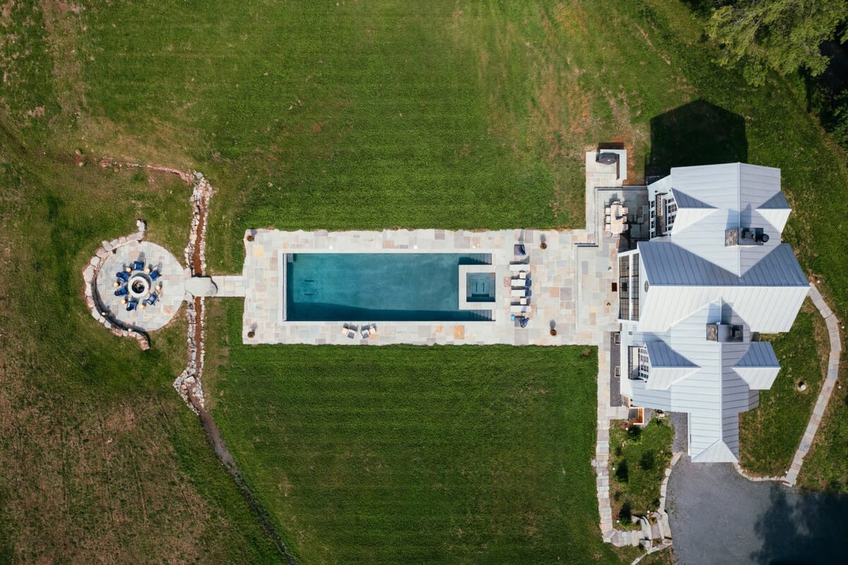 A bird's-eye view reveals the property's layout, featuring the white modern farmhouse, a long rectangular swimming pool, and a circular stone fire pit area, all connected by stone pathways amidst a sprawling green lawn.