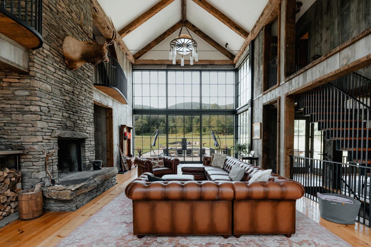 Grand living room with a vaulted ceiling, exposed wooden beams, and a towering stone fireplace. Large black-framed windows offer panoramic views of the outdoor pool and mountains, complemented by brown leather seating.