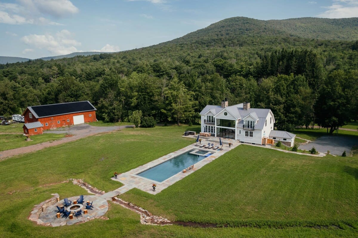 An aerial view showcases the expansive property, featuring a modern farmhouse with a long swimming pool, a circular stone fire pit area, and a large red barn, all nestled within vast green fields and surrounded by forested mountains.