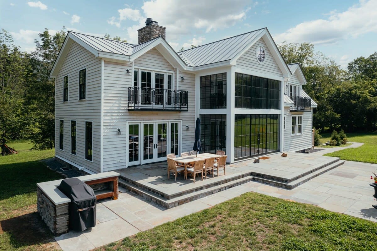The back of the modern farmhouse features a multi-level stone patio with an outdoor dining table and chairs, a built-in BBQ grill station, and large glass doors leading into the house, surrounded by a manicured lawn.