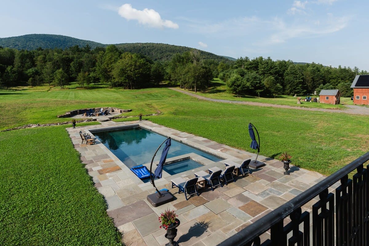 Elevated view of the rectangular swimming pool and surrounding bluestone patio, with lounge chairs and umbrellas. Beyond the pool, a vast green lawn stretches towards distant mountains and a red barn.