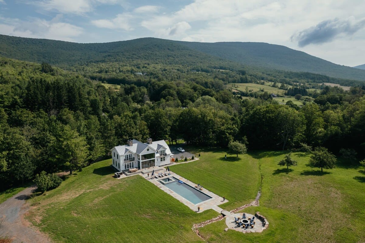 High aerial view showcasing the white farmhouse nestled in a sprawling green valley, surrounded by dense forests and majestic mountains under a partly cloudy sky. The pool and fire pit area are visible.