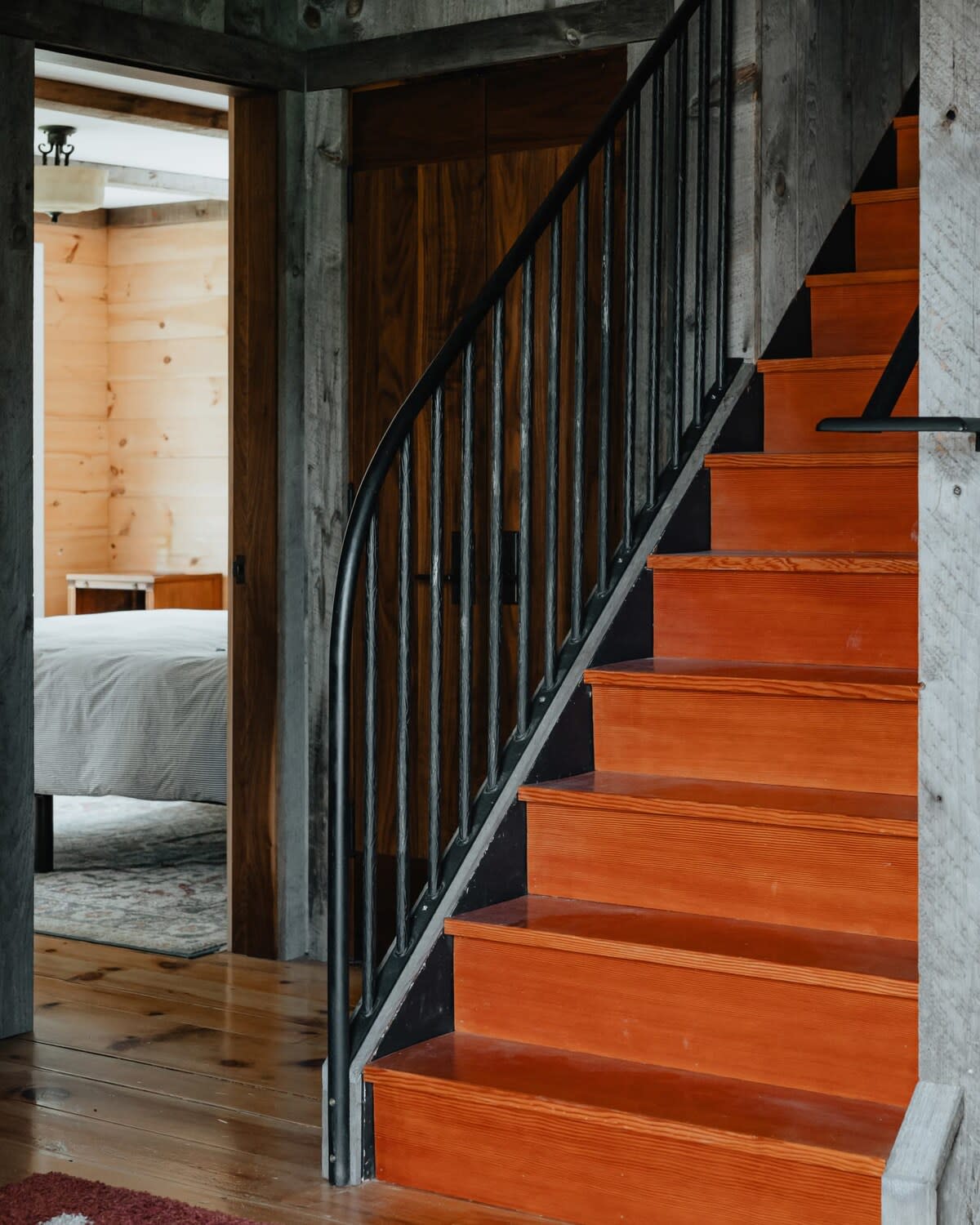 A view of a wooden staircase with dark metal balusters and a curved handrail, leading up from a rustic entryway. The walls are clad in wood planks, and a glimpse of a bedroom is visible through an open doorway.