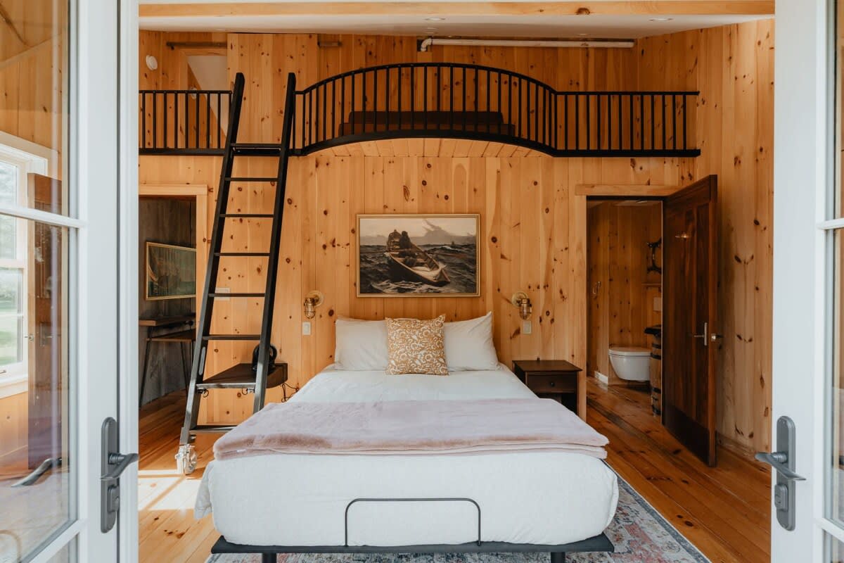 Cozy bedroom with light wood paneling, a queen bed, a black metal ladder leading to a loft, and a painting of a boat above the bed, featuring a rustic cabin aesthetic.