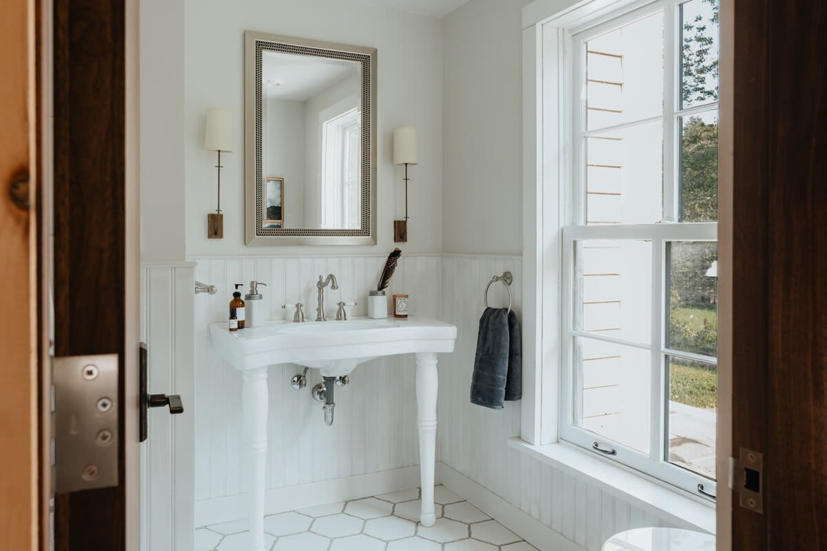 A bright bathroom with white beadboard wainscoting and hexagonal tile flooring, featuring a classic white pedestal sink with a silver framed mirror above. Wall sconces flank the mirror, and a large window provides natural light.