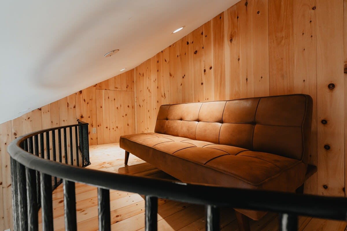 Loft area with light wood paneling, a comfortable brown leather futon, and a black metal railing overlooking the room below, providing an additional lounge or sleeping space.