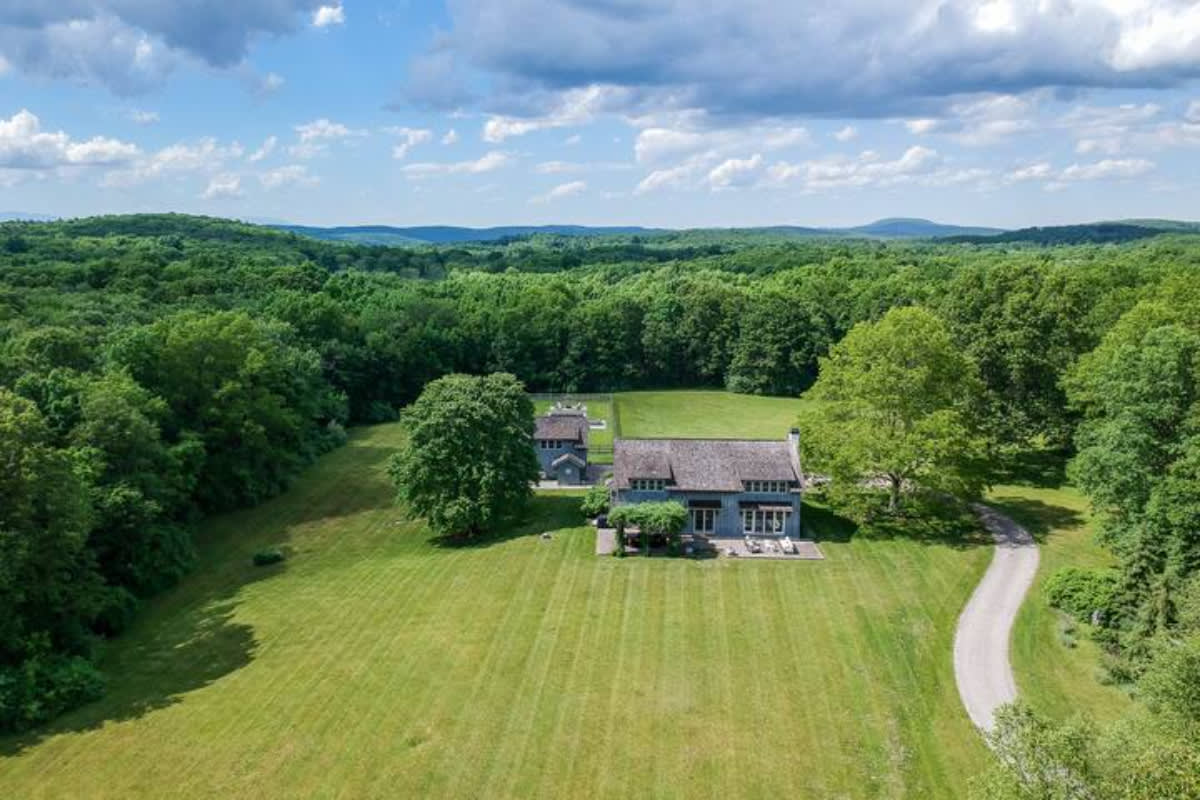 A wide aerial shot displays a light blue house and a separate outbuilding nestled within a vast green landscape, surrounded by dense forest under a partly cloudy sky. A large, open lawn stretches out in front of the house, with a winding driveway and distant rolling hills.