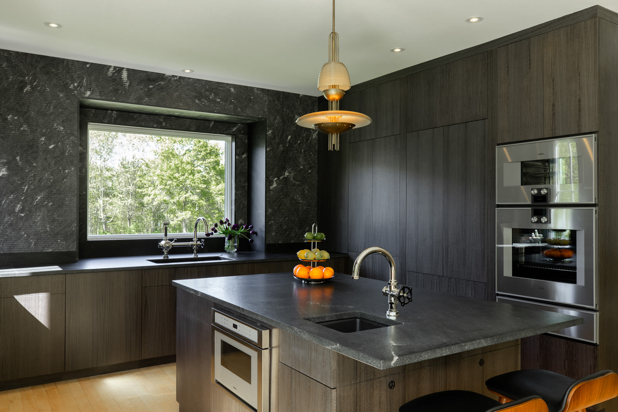 This kitchen view highlights the dark stone island with an integrated sink and microwave, complemented by dark wood cabinetry and black stone countertops. A large window above the main sink offers a view of the outdoors, and a stylish pendant light illuminates the island.