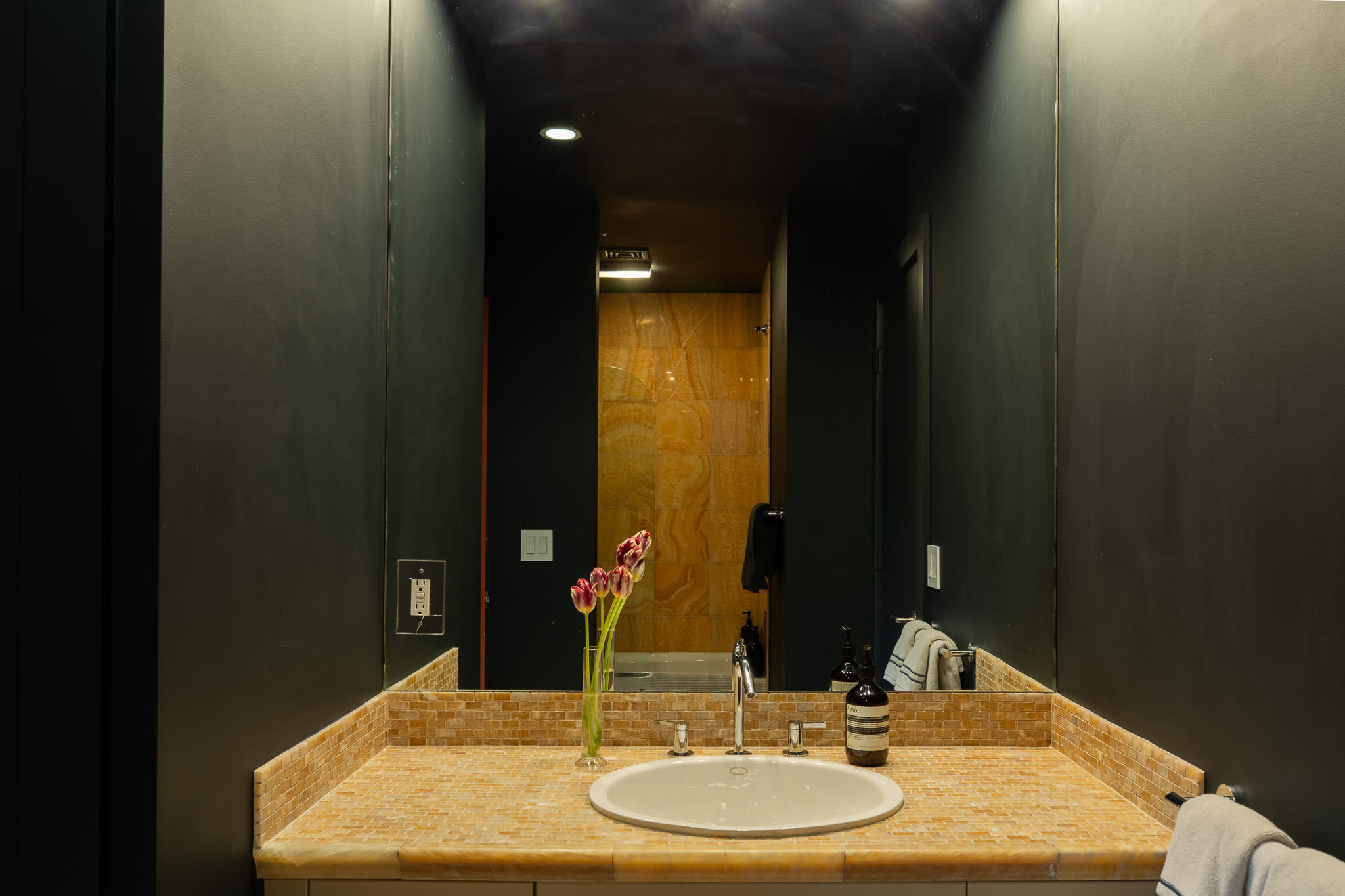 Dark-walled bathroom with a light-colored vanity featuring a white oval sink and a mosaic tile countertop, reflecting a shower area with orange-toned tiled walls.