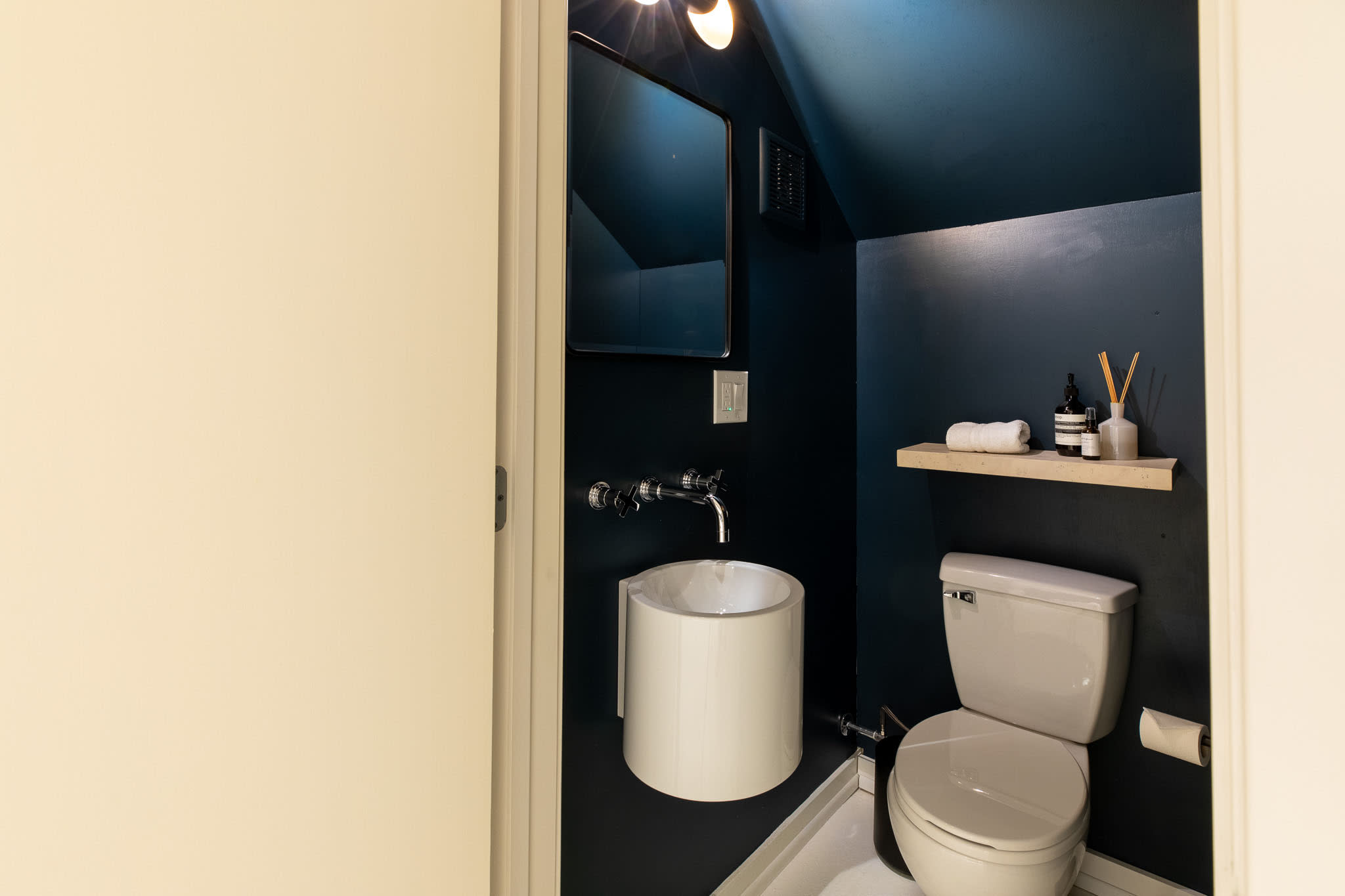 Compact and modern bathroom with dark blue walls and ceiling, featuring a unique cylindrical pedestal sink with a wall-mounted faucet. A toilet is visible next to a small wooden shelf with toiletries.
