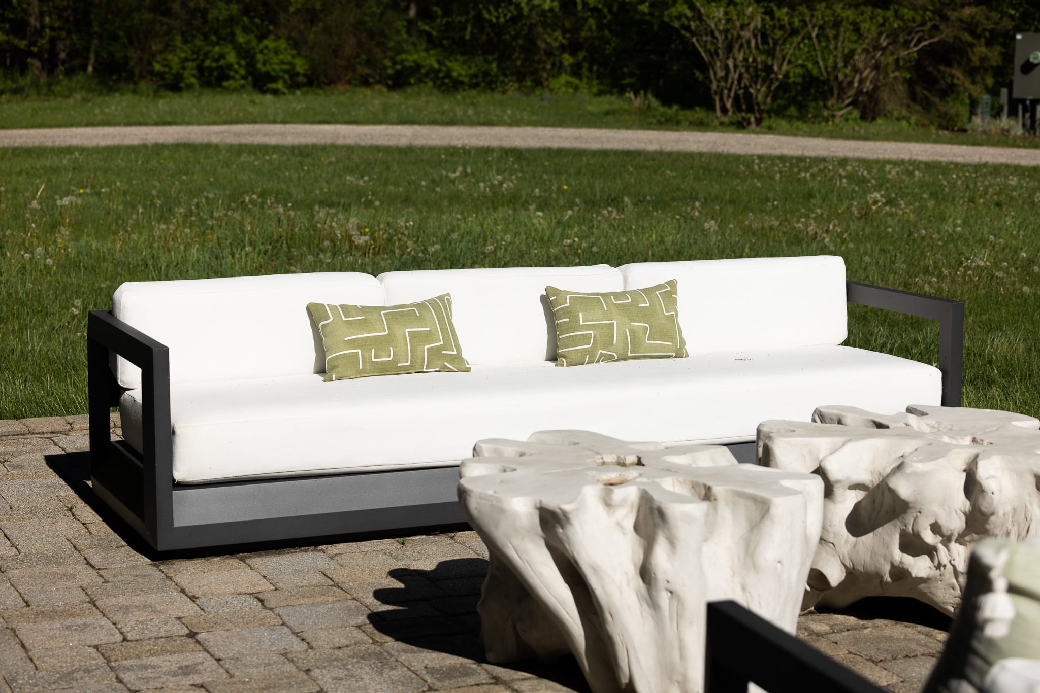 A modern outdoor sofa with white cushions and two green patterned throw pillows sits on a light paver patio, facing a pair of organic-shaped white coffee tables. The sofa has a dark, sleek frame, and the background shows a vibrant green lawn and distant trees under bright sunlight.