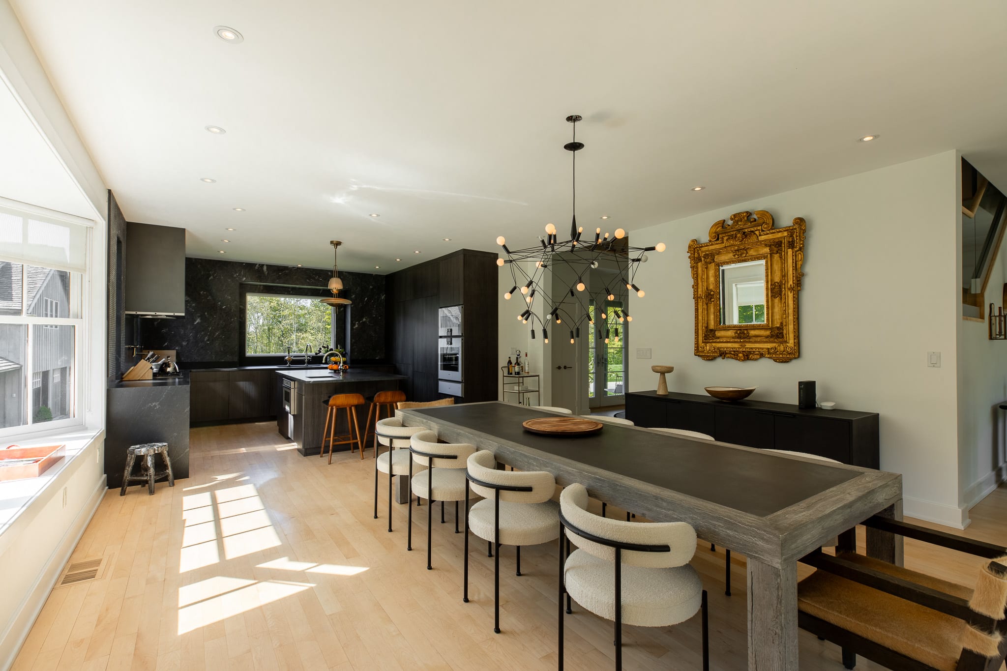 Expansive open-concept space showing the kitchen, dining area, and a large bay window. The kitchen has dark cabinetry and a large island, while the dining area features a long table and modern chairs.