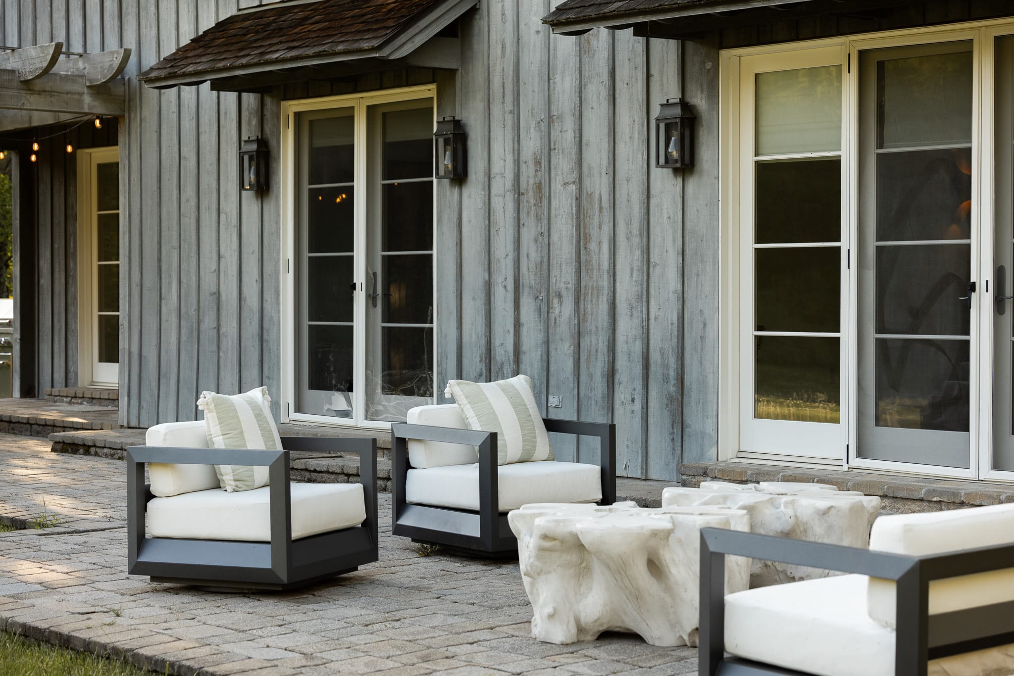 A cozy outdoor patio area features two modern armchairs with white cushions and striped green pillows, arranged around a unique, organic-shaped white coffee table. The chairs have dark frames and are set on a light-colored paver patio next to a weathered gray wooden house with large glass doors.