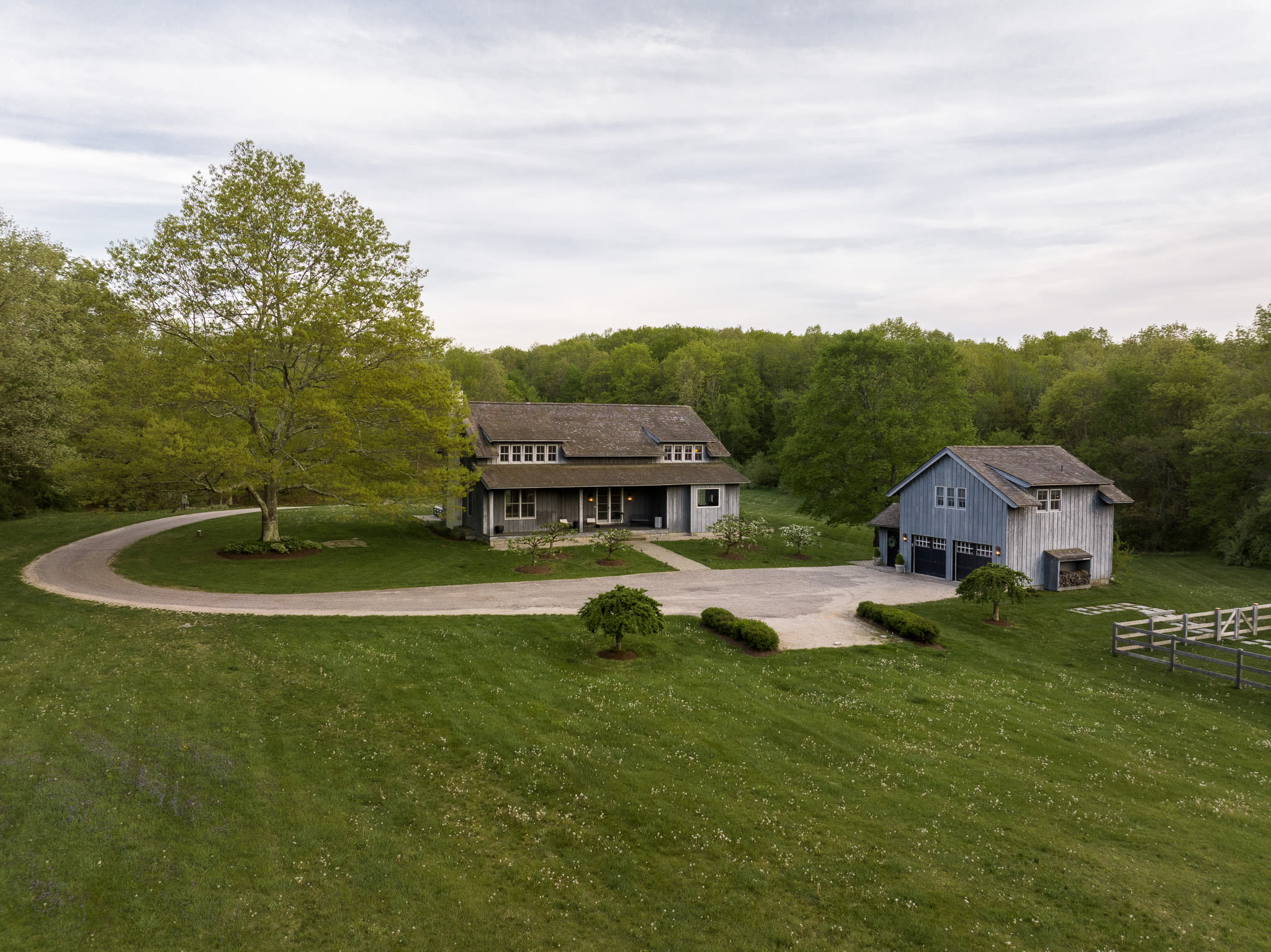 An aerial perspective reveals a light blue main house with a dark roof and a matching detached garage building, both surrounded by a manicured green lawn. A circular gravel driveway connects the structures, with mature trees providing shade and a wooden fence visible to the right.