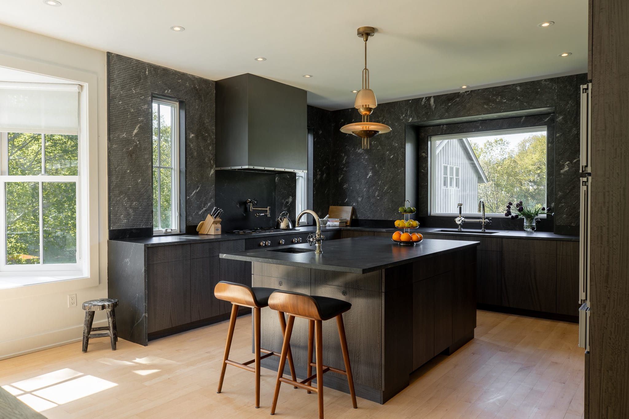 A modern kitchen features dark wood cabinetry and black stone countertops, with a large island providing additional workspace and seating. Stainless steel appliances, including a range hood and double ovens, are integrated into the design, and a decorative pendant light hangs above the island.
