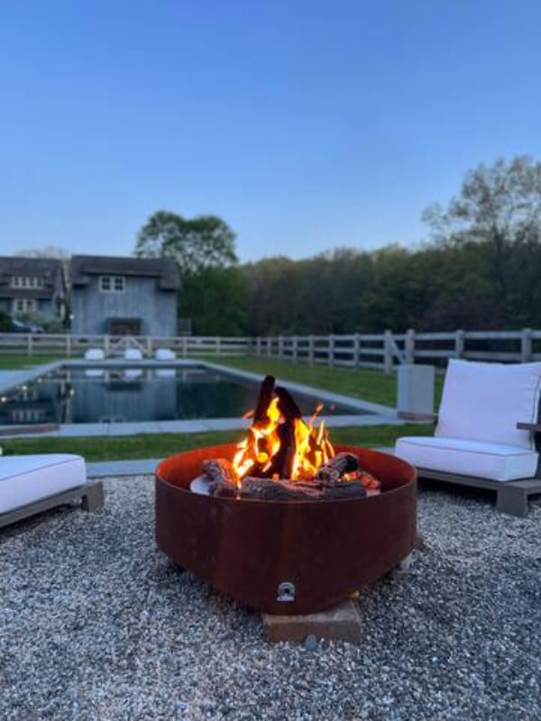 Inviting outdoor fire pit area with a large, rustic metal fire bowl actively burning, surrounded by comfortable outdoor chairs with white cushions. The pool and a distant building are visible in the background under a twilight sky.