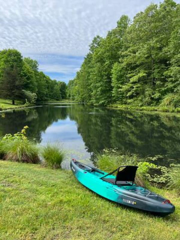 A vibrant teal kayak rests on a grassy bank beside a serene, tree-lined pond or lake, reflecting the lush green foliage and a partly cloudy sky. The water is calm, surrounded by dense woods, suggesting a peaceful natural setting.