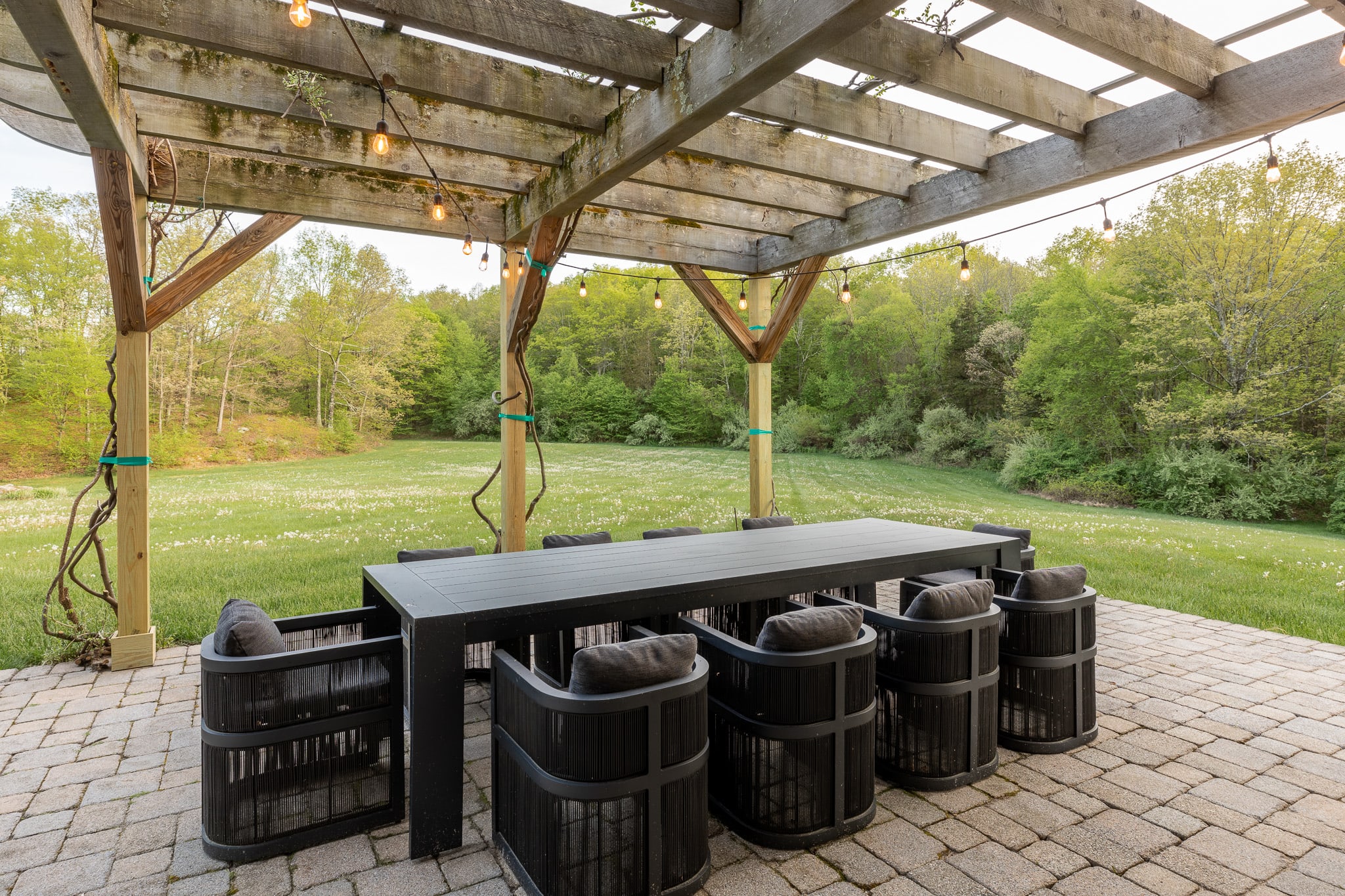 A large outdoor dining area under a wooden pergola, adorned with string lights, features a long black dining table surrounded by eight black woven chairs with dark cushions. The paver patio overlooks a vast, sunny green lawn and a dense forest in the distance.