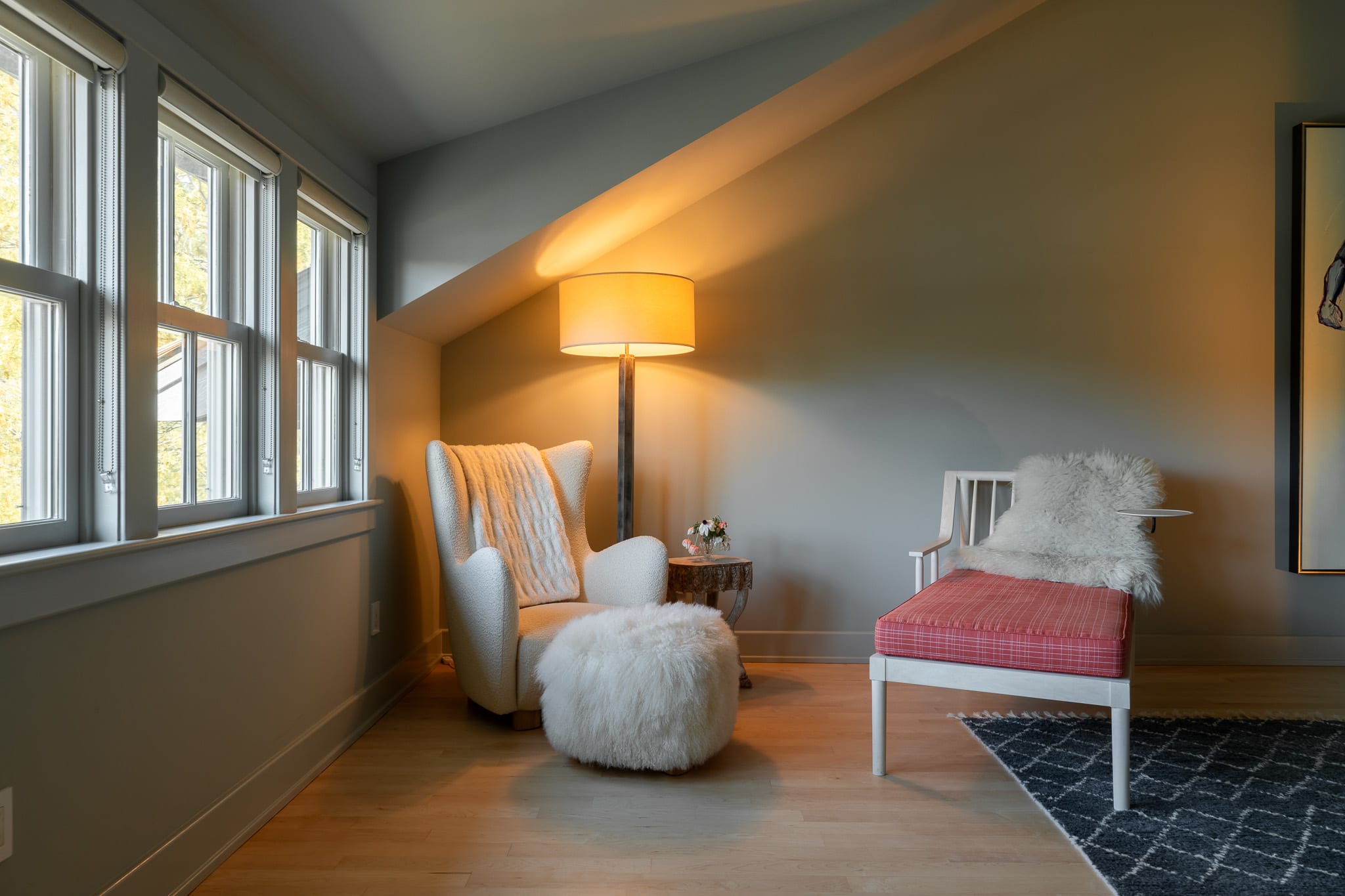 Cozy sitting area in an attic bedroom with a white armchair, ottoman, and a red bench, illuminated by a tall floor lamp and natural light from multiple windows.